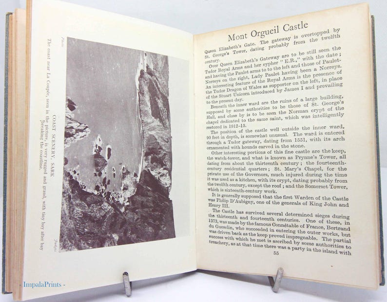 May include: An open book with text and a black and white photograph of a coastal landscape. The text describes Mont Orgueil Castle and coastal scenery on Sark. The book is propped open with a metal book stand.