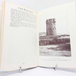 May include: An open book with a sepia-toned photograph of a stone tower on the right page. The left page has text titled "C&acirc;tel & Its Menhir". The book is propped open with a metal book stand.