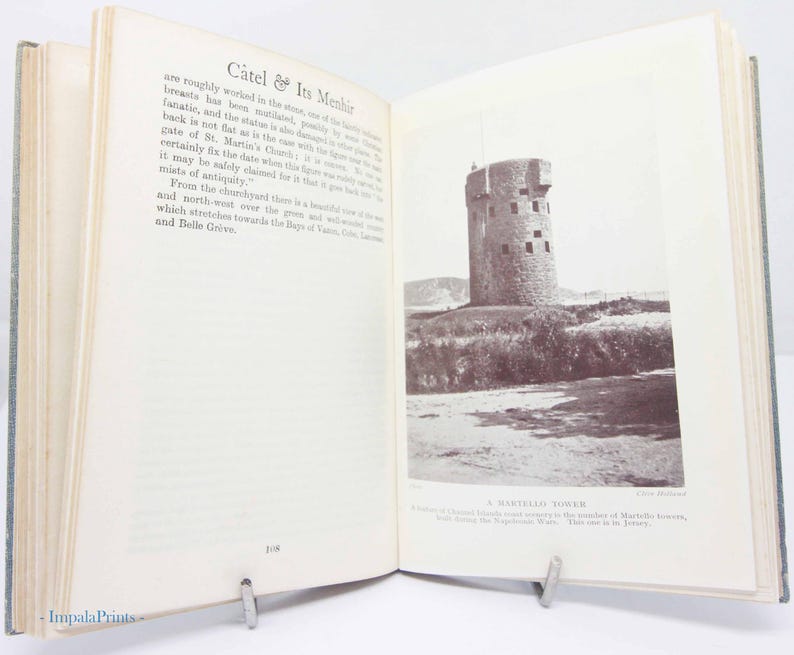 May include: An open book with a sepia-toned photograph of a stone tower on the right page. The left page has text titled "C&acirc;tel & Its Menhir". The book is propped open with a metal book stand.