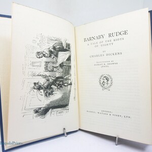 Pode incluir: Um livro de capa dura aberto intitulado Barnaby Rudge: A Tale of the Riots of 'Eighty de Charles Dickens. A página esquerda apresenta uma ilustração a tinta preta e branca de figuras numa sala, enquanto a direita mostra o título.