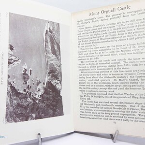 May include: Open book with a black and white photograph of a coastal landscape and text. The text describes Mont Orgueil Castle, including its history and features. The book is propped open with a metal stand.
