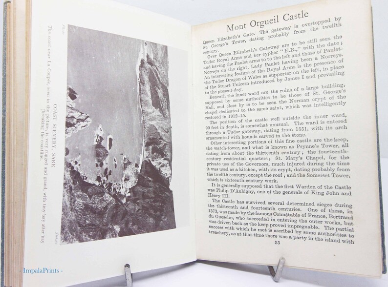 May include: Open book with a black and white photograph of a coastal landscape and text. The text describes Mont Orgueil Castle, including its history and features. The book is propped open with a metal stand.
