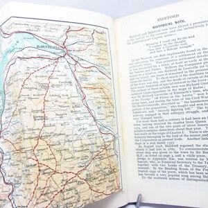 Puede incluir: Libro abierto con un mapa detallado de una región costera, con líneas rojas que indican carreteras y nombres de lugares. La página opuesta contiene texto histórico sobre Bideford, titulado "HISTORICAL NOTE."