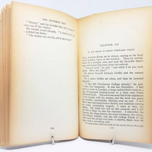Op de afbeelding: Een open boek met verouderde, vergeelde pagina's, opengehouden door een metalen clip. De tekst op de linkerpagina luidt "THE INVISIBLE MAN" en op de rechterpagina "CHAPTER XX". Het boek heeft een groene rug.