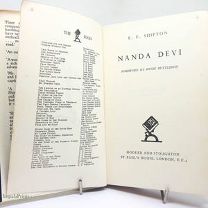 May include: Open book titled "Nanda Devi" by E.E. Shipton, published by Hodder and Stoughton. The book's pages are aged, with a list of other books and authors on the left page. The cover is dark blue.