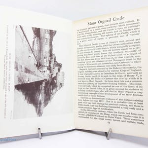 May include: Open book with a black and white photo of Mont Orgueil Castle, Jersey. Text on the right describes the castle's history and features. The book is held open by a metal book stand.