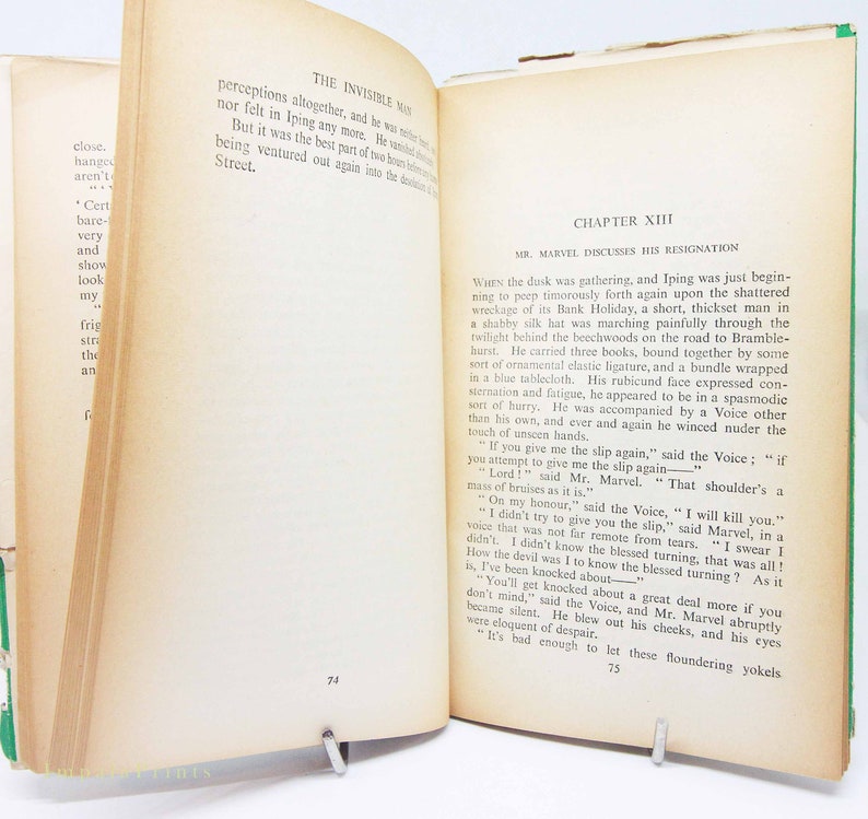 Op de afbeelding: Open boek met tekst uit "De onzichtbare man" van H.G. Wells. De pagina's zijn vergeeld met zichtbare tekst. Het boek wordt opengehouden door een metalen boekensteun. De kaft van het boek is groen.