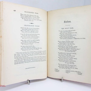 Pode incluir: Um livro antigo aberto com p&aacute;ginas cor de creme e texto impresso em preto. A p&aacute;gina esquerda exibe o poema Craigie-Burn Wood, enquanto a p&aacute;gina direita mostra o t&iacute;tulo Satires e o poema The Holy Fair. O livro tem uma lombada vermelha.