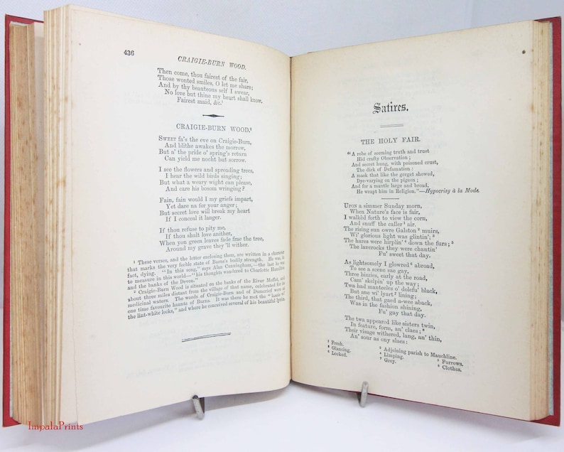 Pode incluir: Um livro antigo aberto com p&aacute;ginas cor de creme e texto impresso em preto. A p&aacute;gina esquerda exibe o poema Craigie-Burn Wood, enquanto a p&aacute;gina direita mostra o t&iacute;tulo Satires e o poema The Holy Fair. O livro tem uma lombada vermelha.