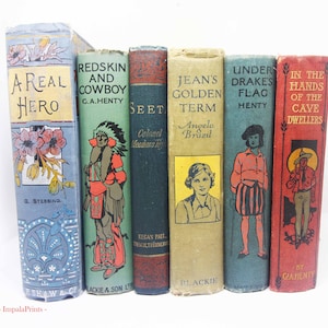 May include: A collection of six vintage hardcover books standing upright. The spines feature various colours including blue, green, dark teal, tan, and red, with illustrated covers and titles such as A Real Hero, Redskin and Cowboy, and In the Hands of the Cave Dwellers.