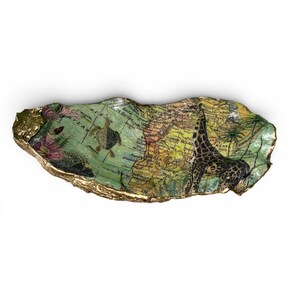 May include: An oyster shell trinket dish with a map design featuring a giraffe and turtle. The dish has a gold-coloured rim and a green and yellow background. The map shows the African continent.