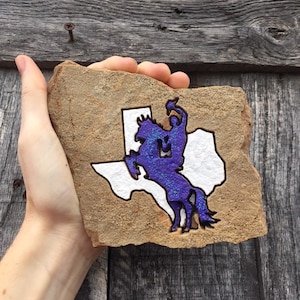 May include: A hand holds a brown rock with a white outline of the state of Texas and a purple silhouette of a cowboy riding a horse.