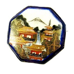 Op de afbeelding: Achtkantige porseleinen broche met een blauwe achtergrond en een geschilderd tafereel van een Japans landschap met een berg, bomen en gebouwen. De gebouwen hebben gouden daken en rode accenten.