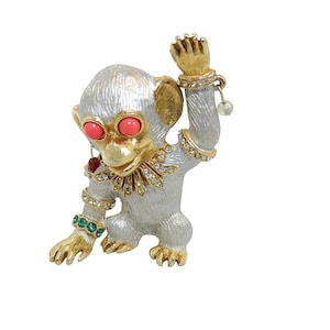 Peut inclure: Une broche en forme de singe argentée et dorée avec des yeux rouges et des bijoux verts et rouges. Le singe porte un collier orné de bijoux et a le bras levé.