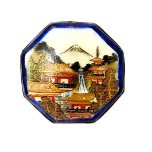 Op de afbeelding: Achtkantige keramische knoop met een blauwe rand en een geschilderd tafereel van een Japans dorp met een berg op de achtergrond.