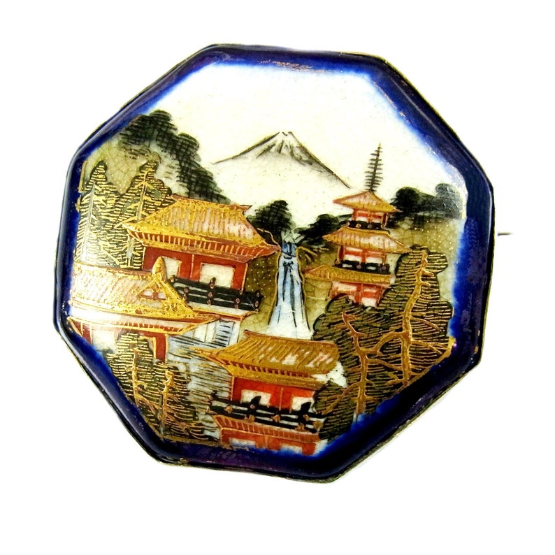 Op de afbeelding: Achtkantige porseleinen broche met een blauwe achtergrond en een geschilderd tafereel van een Japans landschap met een berg, bomen en gebouwen. De gebouwen hebben gouden daken en rode accenten.