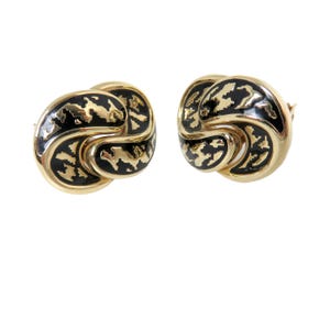 May include: A pair of gold-tone stud earrings featuring a swirling, knot-like design. Each earring has a polished gold metal border and a recessed center filled with black enamel that displays an irregular, abstract gold pattern throughout the surface.