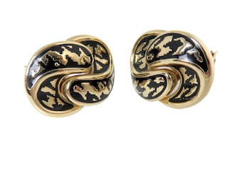 May include: A pair of gold-tone stud earrings featuring a swirling, knot-like design. Each earring has a polished gold metal border and a recessed center filled with black enamel that displays an irregular, abstract gold pattern throughout the surface.