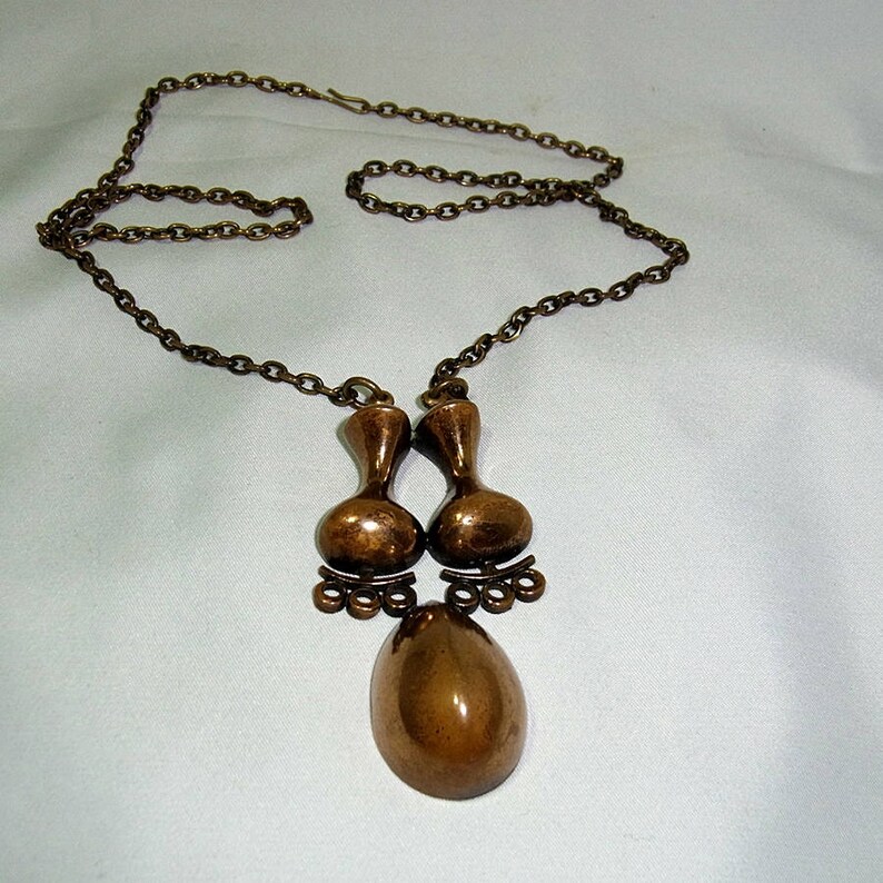 May include: A bronze necklace with a pendant featuring two teardrop-shaped charms and a larger oval-shaped charm.