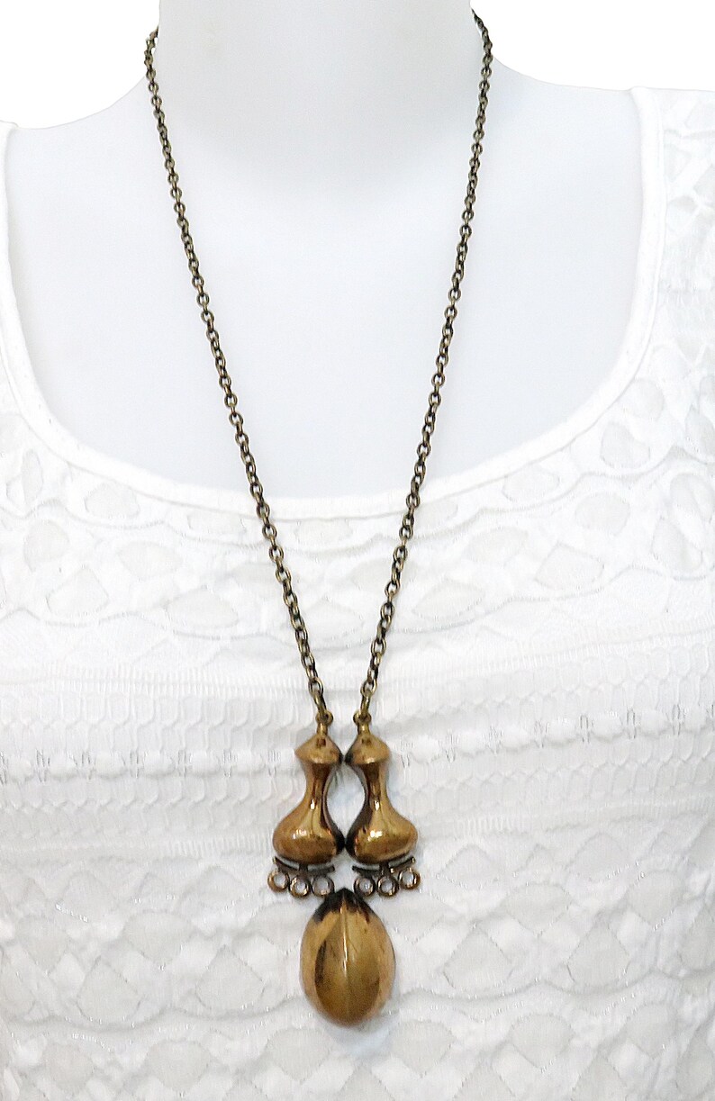 May include: A gold-toned necklace with a unique pendant featuring two teardrop-shaped pieces connected by a smaller teardrop. The pendant hangs from a delicate chain.