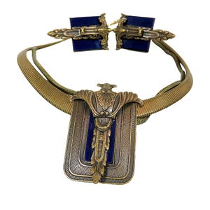 May include: A gold-toned necklace with a blue enamel pendant. The pendant features a stylized design with a central motif and a rectangular frame. The necklace is made of a braided chain.