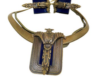 May include: A gold-toned necklace with a blue enamel pendant. The pendant features a stylized design with a central motif and a rectangular frame. The necklace is made of a braided chain.