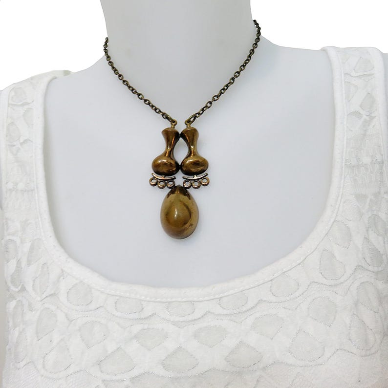May include: A gold-toned necklace with a unique pendant featuring two teardrop-shaped pieces connected by a smaller teardrop. The pendant has a textured, organic look.