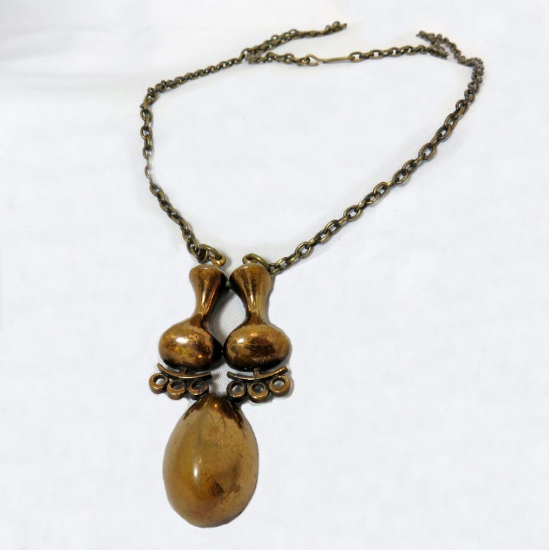May include: A gold-toned necklace with a pendant featuring two teardrop-shaped charms and a larger oval-shaped charm.