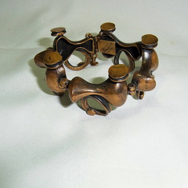May include: A bronze bracelet with a unique, abstract design. The bracelet is made of multiple interconnected pieces, each with a rounded shape. The bracelet has a hinged clasp.