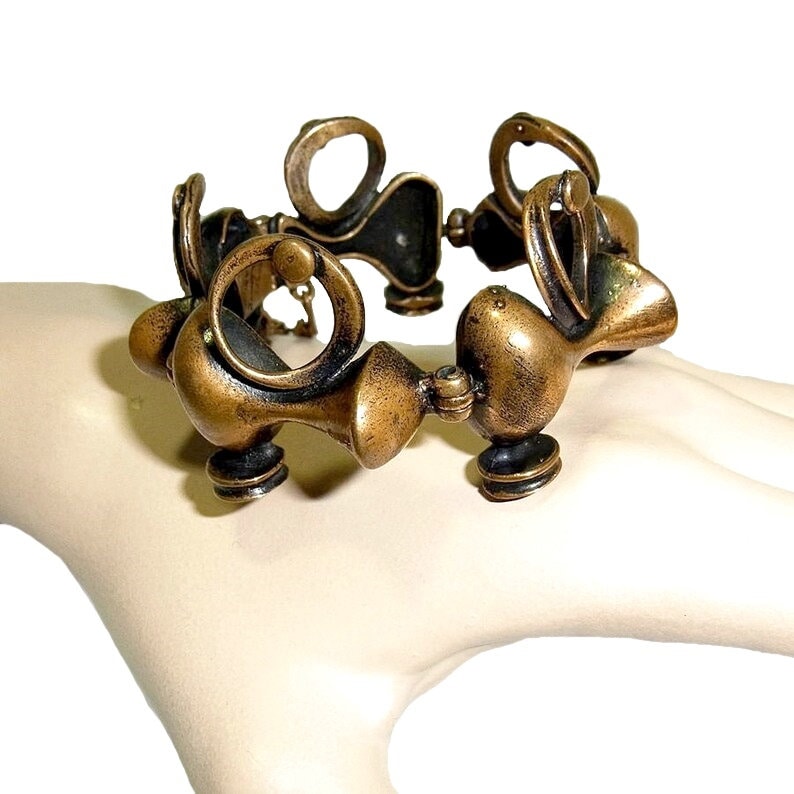 May include: A gold-toned metal bracelet with a unique, abstract design. The bracelet is made up of several interconnected, rounded shapes.