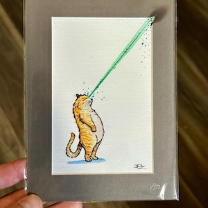 May include: A matted art print featuring an orange tabby cat standing on its hind legs, shooting a bright green laser beam from its eyes toward the upper right corner. The print includes a signature in the bottom right and the text 1/50 in the corner.