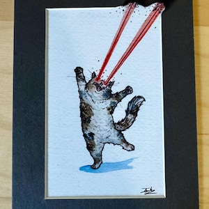 May include: A watercolor illustration of a calico cat standing on its hind legs with its front paws raised. The cat has red laser beams emitting from its eyes, extending upward beyond the top edge of the white paper, framed by a black mat border.