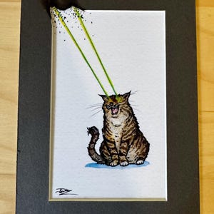 May include: A rectangular matted print featuring a brown tabby cat with its mouth open. Two bright green laser beams shoot from the cat's eyes toward the top left corner, where the paper appears burnt with black speckles. A signature is in the corner.