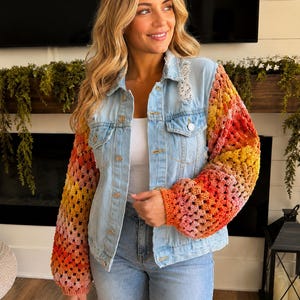 May include: A light blue denim jacket featuring long, loose-knit crochet sleeves in a vibrant gradient of orange, yellow, and pink. The jacket includes a classic collar, metal button closures, two chest pockets, and distressed detailing on the shoulders.