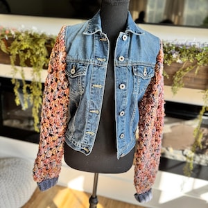May include: A blue denim jacket featuring long sleeves made of multi-colored crocheted yarn in shades of orange, pink, and purple. The jacket includes a classic collar, a front button closure with silver-toned metal buttons, and two chest pockets.