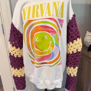 May include: A white crewneck sweatshirt featuring the text NIRVANA in yellow above a colorful, swirling graphic of a smiley face with X eyes. The long sleeves are constructed with alternating horizontal bands of purple and cream crochet.