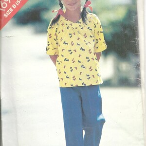 May include: A young girl wearing a yellow short-sleeved shirt with a blue and red print and blue pants. The shirt has a V-neck and the girl has her hair in pigtails with red ribbons.