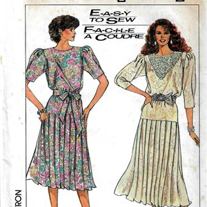 May include: A sewing pattern for a dress with two different styles. The first style is a floral print dress with a gathered skirt and a bow at the waist. The second style is a solid white dress with a gathered skirt and a sash at the waist. The pattern is from Simplicity and the pattern number is 8065.