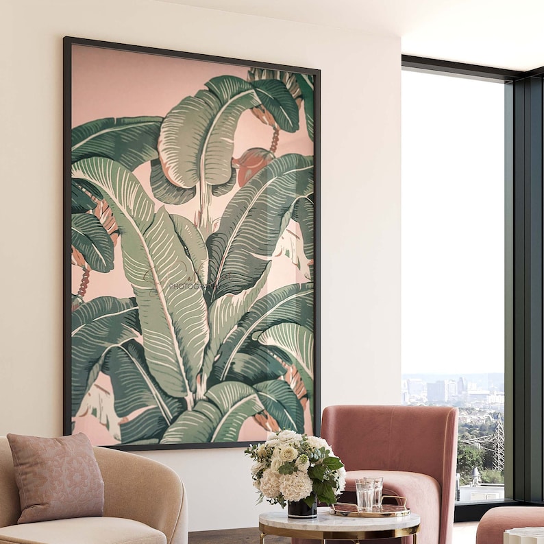 May include: Large framed botanical print featuring green banana leaves against a soft pink background. The leaves have detailed veining and are set in a black frame. The print is displayed in a modern living room setting with a pink armchair and a small table.