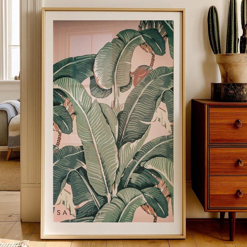 May include: A framed botanical print featuring large green banana leaves with white veins against a soft pink background. The artwork is in a gold frame and includes the text "SAL" in the lower left corner. The print is displayed in a well-lit room.