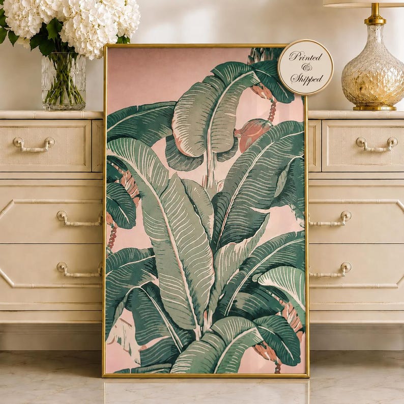 May include: A framed botanical print featuring large green banana leaves against a soft pink background. The leaves have detailed veining and are arranged in a lush, overlapping pattern. The frame is gold-colored, and the print is labeled "Printed & Shipped".