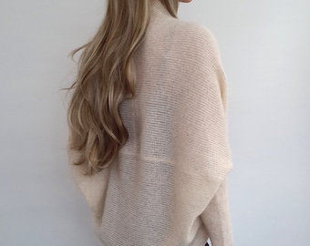 May include: A cream-colored knitted sweater with a loose, draped fit. The sweater has a high neckline and long sleeves. The model is wearing black leggings.