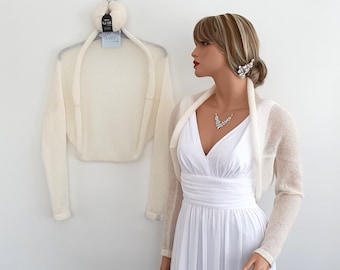 May include: A white knitted shrug with long sleeves. The shrug is draped over a white dress form and a white dress is shown on a mannequin.