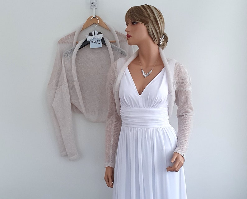 May include: A light beige knitted shrug with long sleeves. The shrug is draped over a white dress form.