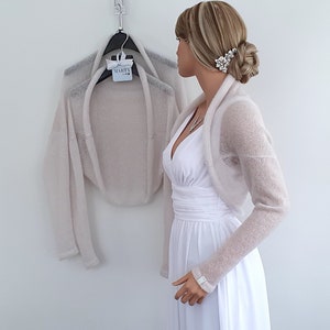 May include: A light beige knitted shrug with long sleeves. The shrug is draped over a white dress form.