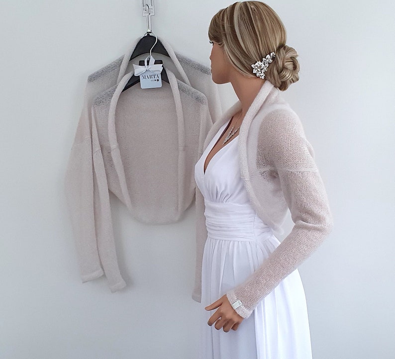 May include: A light beige knitted shrug with long sleeves. The shrug is draped over a white dress form.