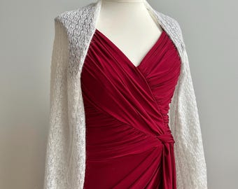 May include: A white knitted shrug with long sleeves and a delicate openwork pattern. The shrug is draped over a red dress with a V-neckline.