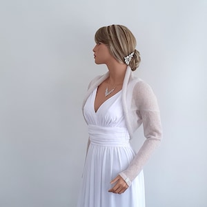 May include: A white knitted shrug with long sleeves. The shrug is made of a lightweight, sheer fabric and has a draped neckline. The shrug is being worn by a mannequin over a white dress.