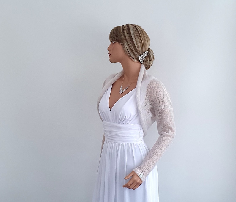 May include: A white knitted shrug with long sleeves. The shrug is made of a lightweight, sheer fabric and has a draped neckline. The shrug is being worn by a mannequin over a white dress.