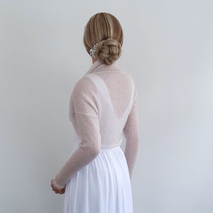May include: A sheer, light pink knitted shrug with long sleeves. The shrug is draped over the shoulders and arms of a person wearing a white dress.